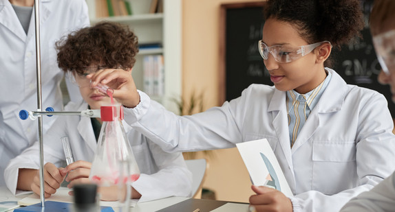 Eine Schülerin und ein Schüler experimentieren im Labor mit weißem Kittel und Schutzbrille